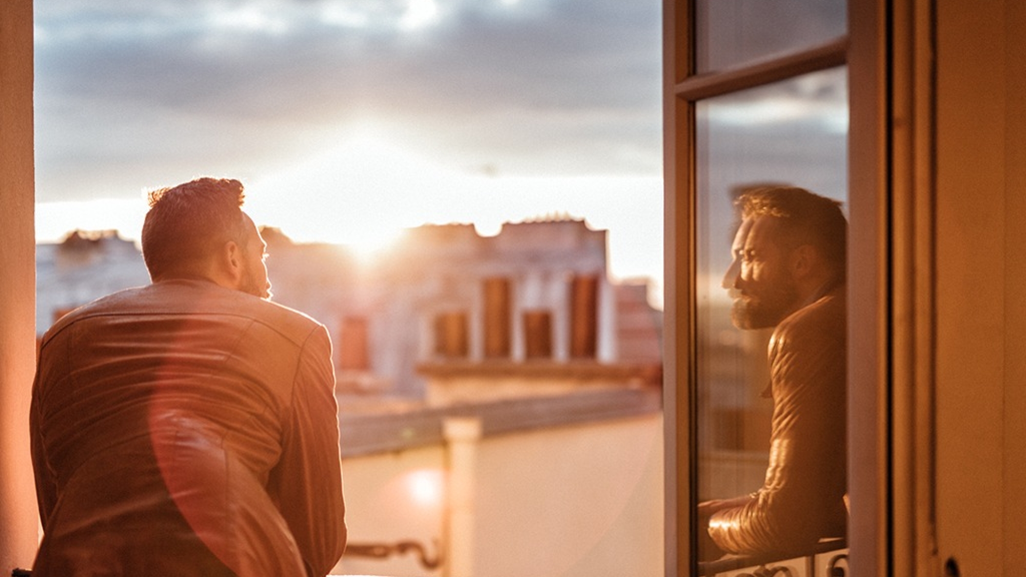 bearded man looking out over the city at sunrise and reflecting himself in window glass